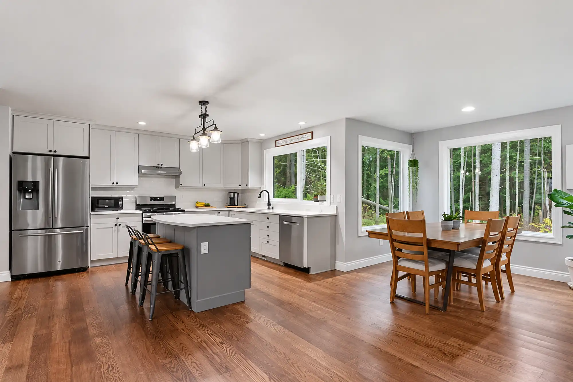 Open kitchen with white cabinets, a gray island, wood flooring, and a dining area beside large windows.