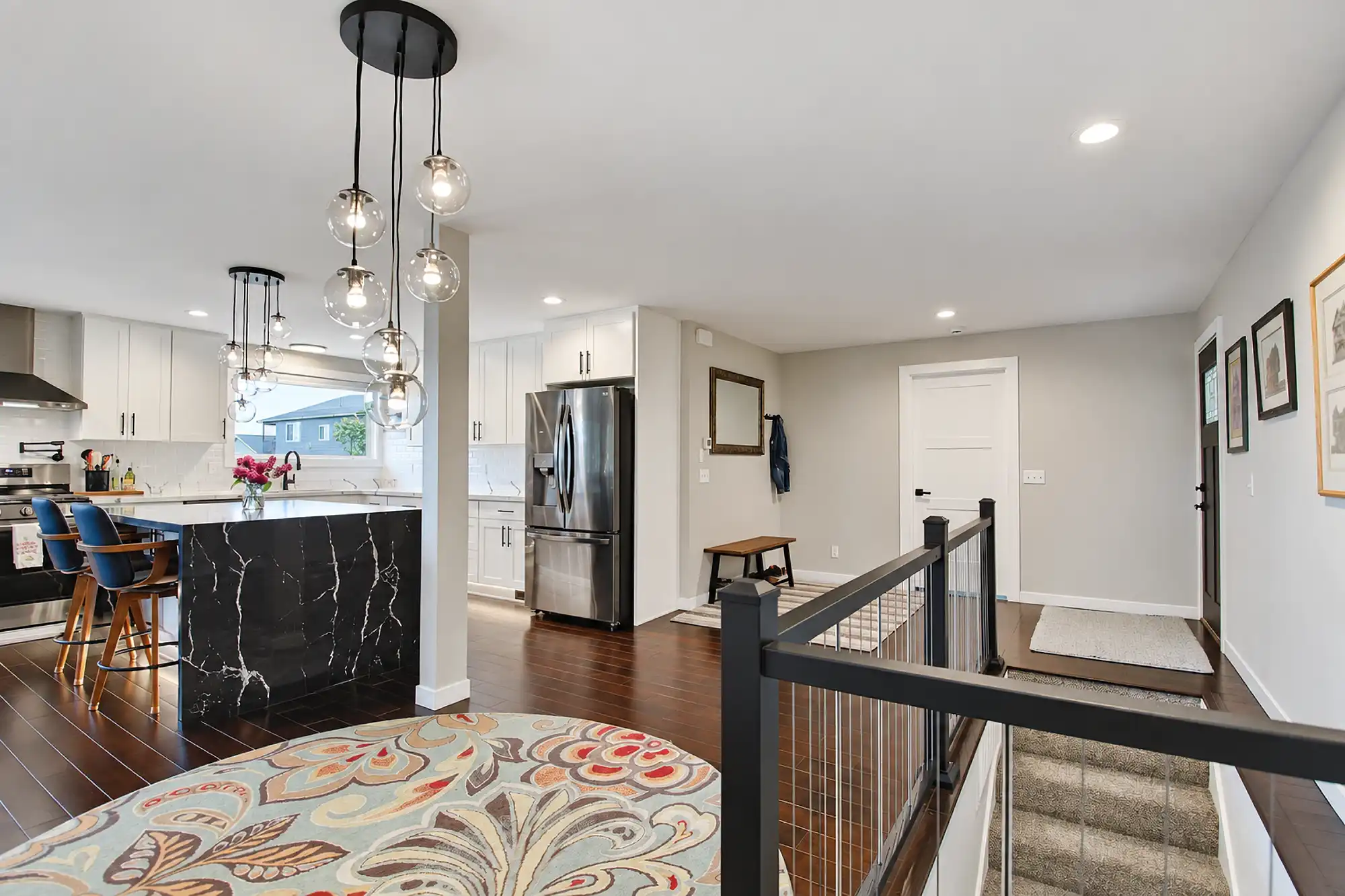 Open kitchen beside a stair landing with dark railings, a black veined island, and glass pendant lights.