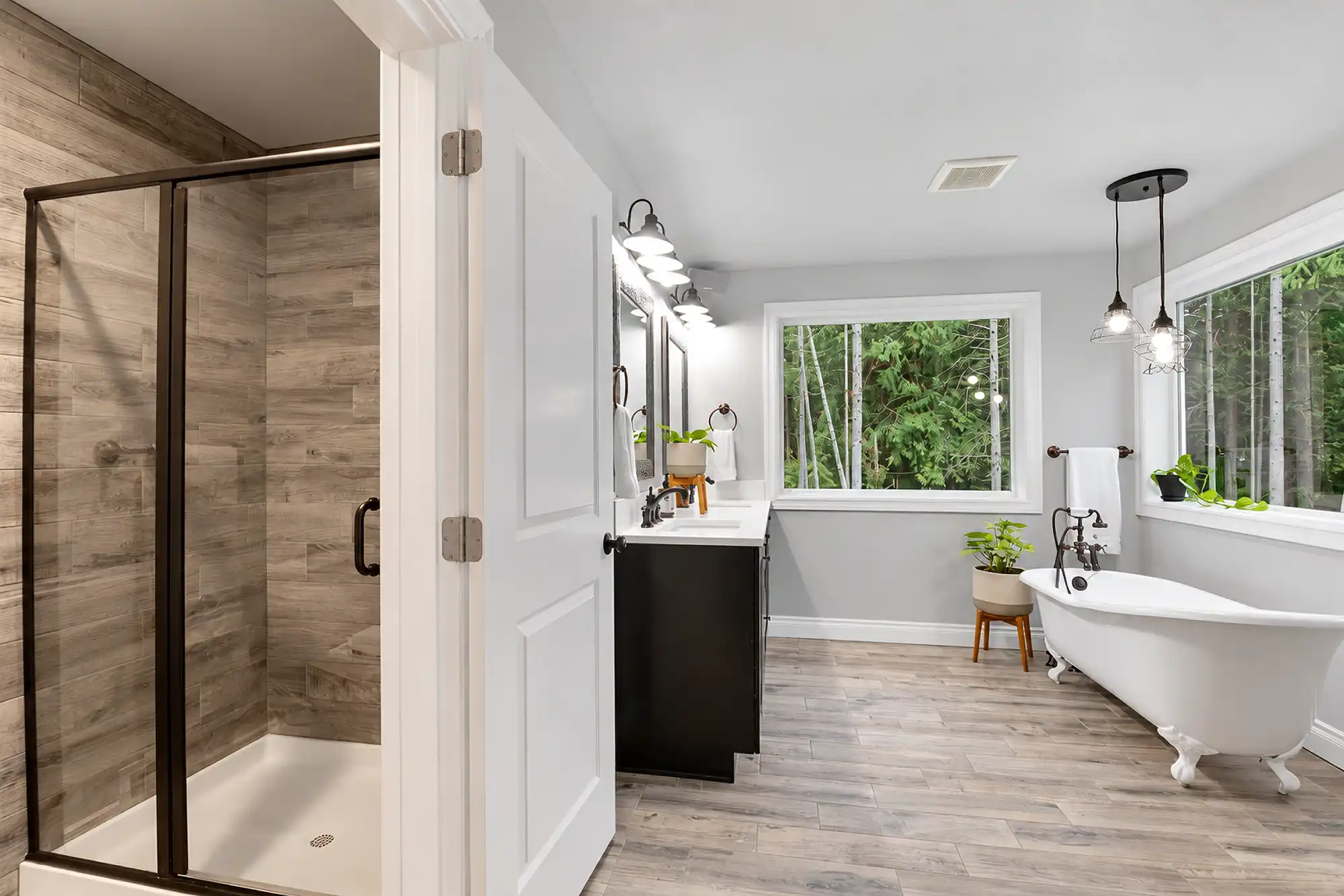Bathroom with a white clawfoot tub, glass-enclosed shower, dark vanity, and large windows overlooking trees.