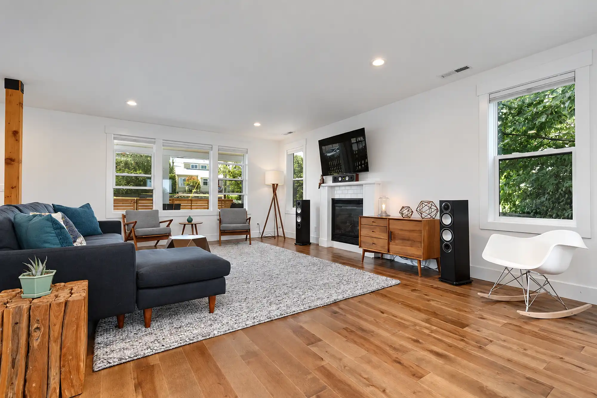 Living room with a dark sectional, wood flooring, large windows, and a fireplace beneath a wall-mounted television.