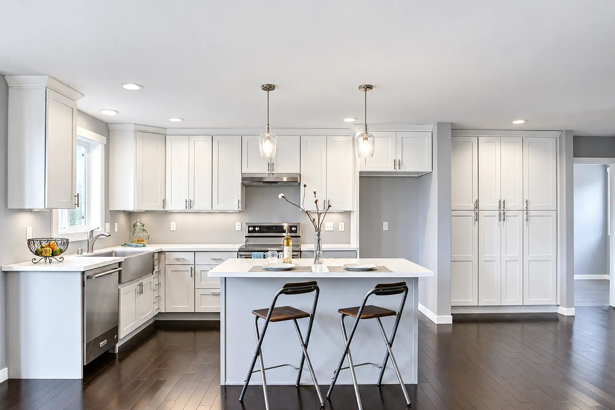 White kitchen with a gray island, two bar stools, pendant lights, and tall pantry cabinets.